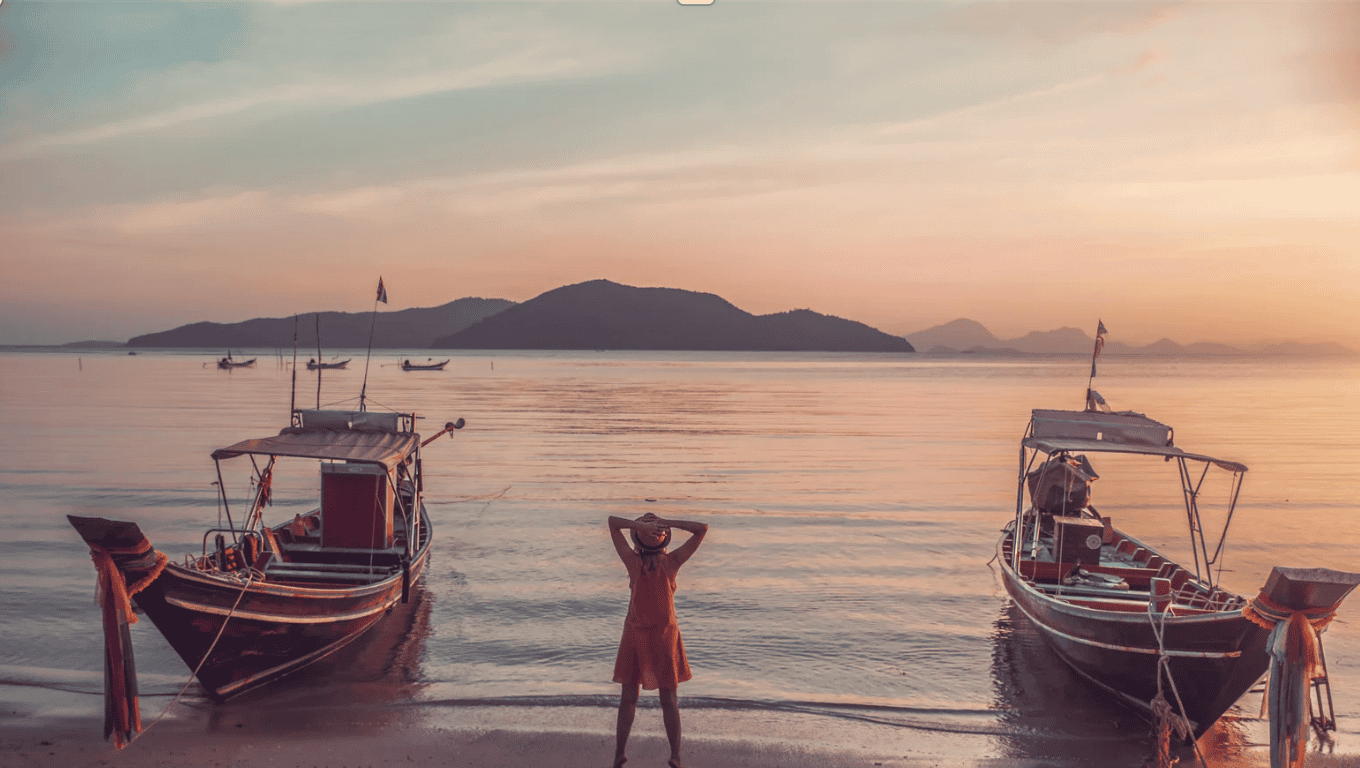 Woman in orange dress between two longtail boats at Thai beach sunset